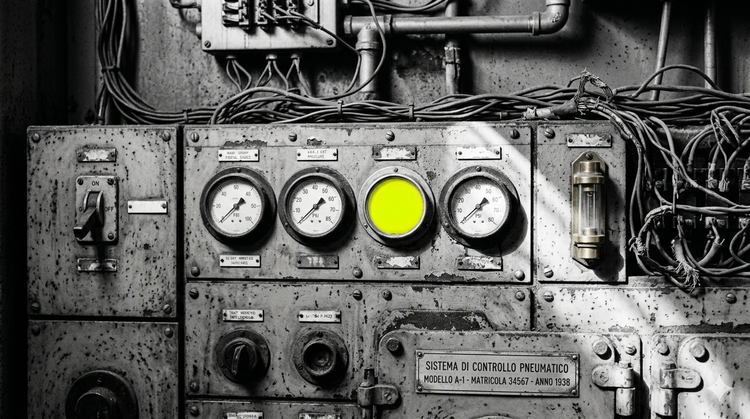 A black and white photograph of a vintage, rusted industrial control panel with numerous wires and four large pressure gauges. One of the circular gauges glows bright, luminous yellow.