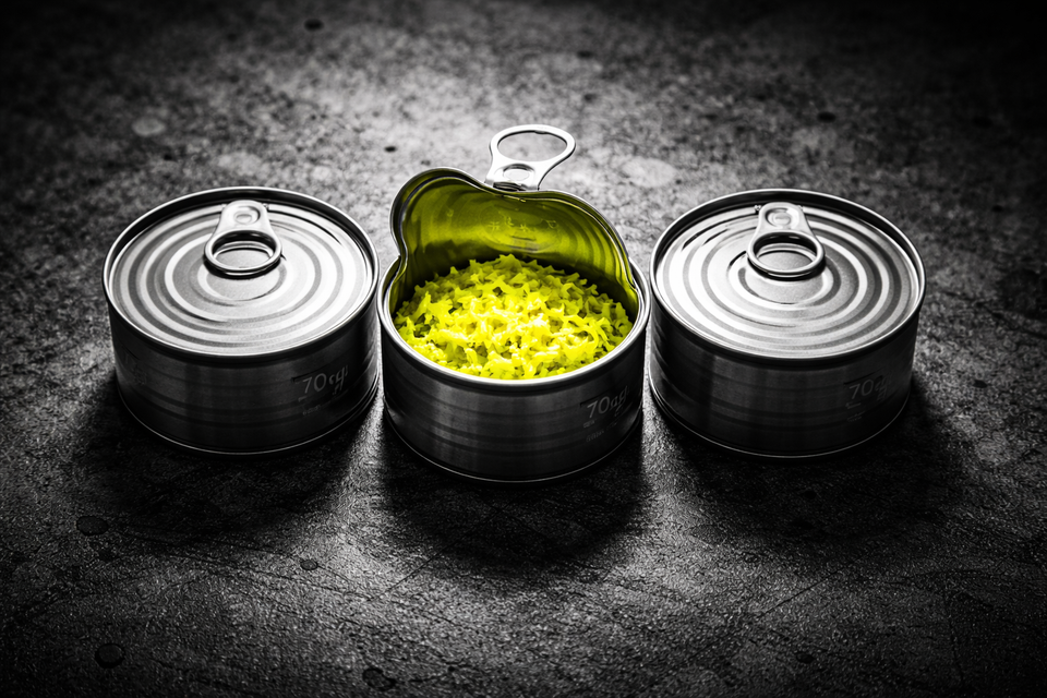 Black-and-white overhead photo of three identical cat food cans on rough concrete; the center can is open, revealing glowing acid-yellow shredded contents under harsh industrial lighting.